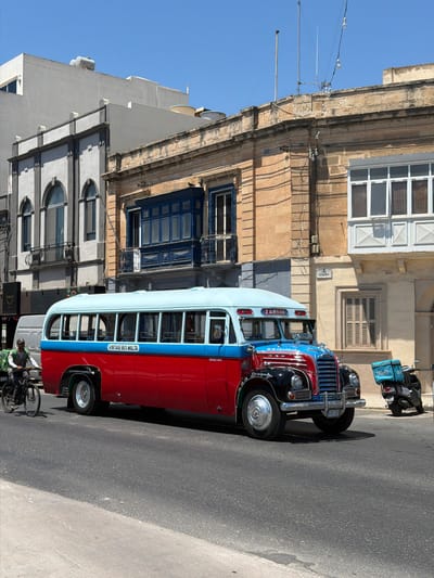 This image captures a classic red and light blue bus, identified by text on its side as "VINTAGE BUS MALTA" and on the front as "ZABBAR," driving down a street in Gżira, Malta. The bus has a chrome grille, round headlights, and appears to be in operational condition. To the left of the bus, a person on a bicycle, wearing a backpack, is riding in the same direction. In the background, there are multi-story buildings with traditional Maltese architecture, including stone facades, balconies, and various window styles. A black scooter with a light blue delivery box labeled "Wolt" is parked to the right of the bus. The scene is set on a clear, sunny day, with the street and sidewalk visible in the foreground.