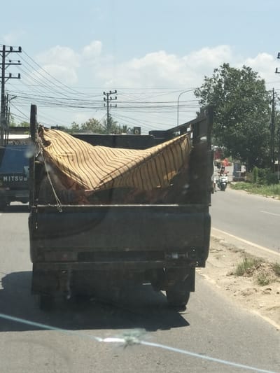 The image captures a scene from the city of Gampongaceh, Indonesia, on a sunny day with a clear blue sky dotted with white clouds. The focal point is the rear of a dark-colored, open-bed truck, likely a pickup truck, driving on a paved road. The truck is loaded with what appears to be bundled goods or possibly a mattress covered with a striped, light-colored fabric. The fabric has a pattern of thin, parallel lines in varying shades of yellow and brown.

To the left of the truck, the side of another vehicle, a blue truck with "MITSU" visible on its side, is partially seen. The road is flanked by a grassy verge on the right and a line of trees and utility poles with numerous wires on the left, characteristic of urban or semi-urban environments. In the distance, a person on a white motorcycle with a red flag is riding on the same road. The overall impression is a typical roadside scene in Indonesia, with vehicles navigating a road amidst infrastructure and vegetation.