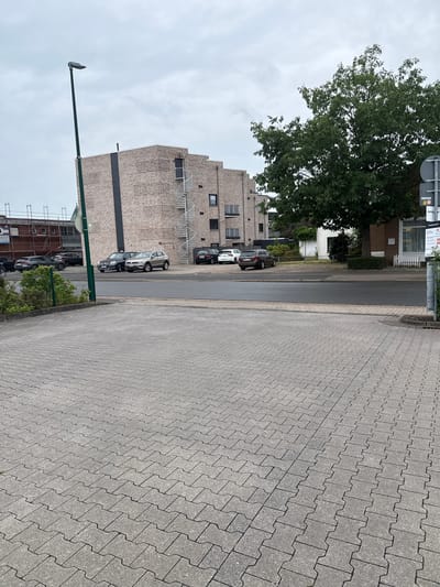 The image depicts an outdoor scene in Sande, Germany, likely in a parking area or a street adjacent to a building. In the foreground, a grey paver sidewalk stretches across the frame, with a repeating pattern of interlocking stones. Behind the sidewalk, a paved road is visible, with several cars parked along its edge.

The cars visible include a dark-colored SUV, a light-colored SUV, and another dark-colored sedan. Further in the background, a modern-looking brick building with spiral staircases attached to its exterior dominates the scene. The building appears to be several stories high. To the right of the building, a large, leafy green tree stands prominently.

A tall street lamp is positioned to the left of the building. The sky above is overcast with grey clouds. The environment appears to be a mix of urban and green space, suggesting a mixed-use area within the city. No people are visible in the image, and no actions or interactions are taking place. The overall impression is one of a calm, everyday scene.