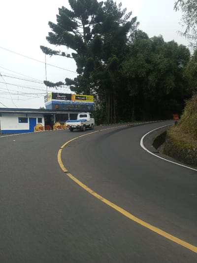 The image displays a curved asphalt road in what appears to be a rural or mountainous setting, under an overcast sky. The road features a solid yellow line indicating the center or lane division on the left side of the curve, and a white solid line marking the shoulder on the right side.
To the left of the road, there is a single-story building with white walls and blue trim around its windows and doors, topped with a corrugated metal roof. In front of this building, a small white pickup truck with a blue tarp covering its bed is parked. Decorative or barrier-like elements, which appear to be yellow and red barrel or cone-shaped objects, are placed along the front of the building.
Above the building, there are two large black signs with yellow lettering. One clearly reads "Alkaline" and another word that appears similar to "Amalline". Below these, a prominent blue sign with white text is visible, which reads: "POS 6 B" and "SATLANTAS POLRES BATUR". This indicates it is Traffic Police Post 6 B for the Batur Resort Police. Utility lines and cables are visible crisscrossing the sky above the building.
Behind the building and extending upwards, a tall, dense pine-like tree is visible. To the right of the road, a steep, grassy bank with some visible rocks slopes upwards, and a dense forest of dark green trees, possibly bamboo or other broadleaf trees, borders the road. Further down the road on the right, an orange barrier or cone is partially visible. The overall impression is a quiet roadside scene on a cloudy day.