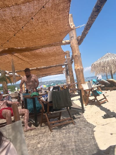 The image depicts a beachside establishment in Kranevo, Bulgaria. A thatched umbrella provides shade over a seating area with wooden tables and chairs set on the sand. In the foreground, a man wearing red shorts is seated, partially obscured by a chair. To his right, a shirtless man with a beard stands near a table laden with drinks, appearing to serve or prepare them. The background reveals a beach with more seating, umbrellas, and the sea beyond, under a clear blue sky. The scene suggests a relaxed, outdoor dining or drinking environment on a sunny day.
