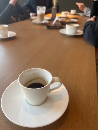 The scene is an indoor setting, likely a cafe or restaurant, located within the city of Rietheim-Weilheim, Germany. A light brown wooden table, appearing to be laminate or light wood, occupies the majority of the frame. In the immediate foreground, a white ceramic espresso cup, containing a small quantity of dark liquid with visible coffee crema residue on its interior walls, rests centered on a round white ceramic saucer.
Further back on the table, blurred and out of focus, are several other items and parts of individuals. To the left, a white ceramic cup on a saucer is partially visible. Centrally, a tall glass containing a light-colored, frothy beverage, likely a latte, is present. Nearby, a dark rectangular object, consistent with a smartphone, lies on the table. A small, dark, cylindrical object with light-colored, vertical decorative elements (possibly dried grasses in a vase) stands between the glass and another white ceramic cup on a saucer to the right. A metallic dish with indeterminate contents is also visible.
At least four individuals are seated around the table, though their faces are obscured or out of frame. To the far left, a person's arm, clad in dark clothing, is partially visible. Behind the tall glass, another individual's torso, also in dark attire, is discernible. To the right, an arm with an extended index finger, suggesting a gesture of pointing or emphasis, is visible, wearing a light-colored sleeve beneath a darker outer garment. Further right, another person's dark sleeve is partially seen. The overall impression is a social gathering with drinks and potential discussion.