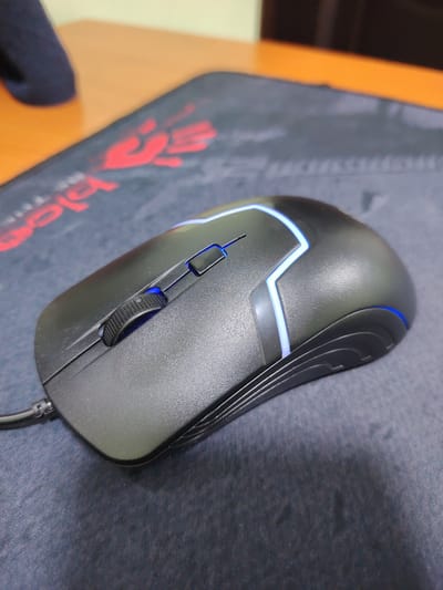 A black wired computer mouse with blue LED accents rests on a black mousepad with a red logo on a wooden desk. The mouse has a textured scroll wheel and two distinct buttons. The mousepad has red lettering and graphic elements. In the background, a dark blue object is partially visible on the desk, and a portion of the wooden desk surface is visible. The lighting is indoor and appears to come from above and to the left of the scene. There is no indication of a city or specific location within Uzbekistan.