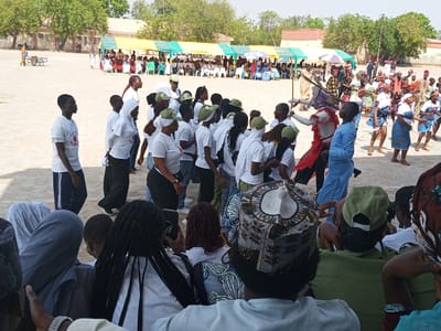 This image depicts an outdoor gathering, likely a cultural or community event, taking place in Ajiwa, Nigeria. The scene is set under a bright, sunny sky, suggesting daytime. In the background, several green and yellow tents are set up, indicating a temporary event space. People are gathered under these tents, some seated on chairs, while others are standing.
In the foreground and mid-ground, a diverse group of people are engaged in various activities. Many individuals are wearing white t-shirts, some with visible text that is difficult to decipher completely but appears to include the word "PLAYERS". A significant portion of the crowd is also wearing green caps.
A notable element of the scene is a group of people in traditional attire, some adorned with beads and colorful fabrics, dancing or moving rhythmically. One individual, dressed in a striking red and purple costume with white face paint, stands out as a central figure in the dancing group, possibly representing a traditional masked dancer or performer. Another individual in a flowing golden robe is also present among the performers.
The overall atmosphere appears lively and celebratory. The ground is a sandy or dusty open area, typical of many outdoor venues in the region. The presence of trees in the background provides some natural element to the setting. The people in the foreground, particularly those closest to the camera, are partially obscured, suggesting they might be observers or participants at the edge of the event. Their expressions are not clearly visible due to their positioning.