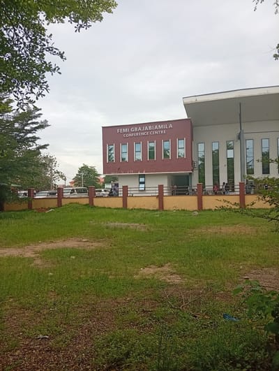 The image depicts the Femi Gbajabiamila Conference Centre in Ijanikin, Nigeria. The conference centre is a two-story building with a maroon section and a white section. The building has several vertical windows. A brown and yellow fence runs along the front of the building. People are visible standing near the entrance. A grassy area is in the foreground, with trees partially obscuring the view on the left and right sides of the frame. Several vehicles are parked behind the fence. The sky is overcast.