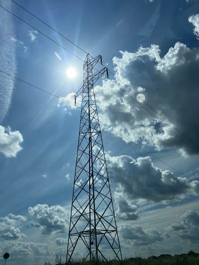 A tall, dark metal power line pylon stands silhouetted against a bright blue sky filled with white and dark grey clouds. The sun is visible, shining brightly and creating a lens flare effect. Several power lines stretch across the sky from the pylon. The pylon is a lattice-like structure, wide at the base and tapering towards the top. Below the pylon, the ground is covered in dry grass and some low-lying vegetation, with a small round sign visible on the left side of the frame. This scene is located in the city of Fasty, Poland.