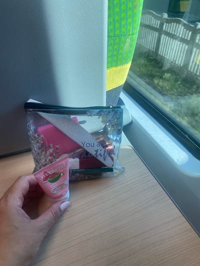 A hand holds a small tube of "weekend feeling" hand cream with a watermelon design in front of a clear makeup bag on a wooden table. The makeup bag contains a pink nail file, lipstick, and other small items. The scene appears to be inside a train car, with a window showing greenery and a fence outside. The train seat fabric is visible on the left side of the frame, featuring a green and yellow pattern. The overall lighting suggests it is daytime.