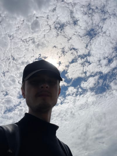 A low-angle, head-and-shoulders selfie is captured against a cloudy sky with the sun partially obscured. The subject, a young person wearing a black baseball cap and a black turtleneck shirt, is looking directly at the camera. Their face is in shadow, with only the lower part of their nose and a faint mustache and goatee visible. The background is dominated by a dense layer of cumulus clouds, interspersed with patches of blue sky. The image appears to be taken outdoors, possibly in Diepenbeek, Belgium, given the context provided. There are no other individuals or objects of significance visible in the frame.