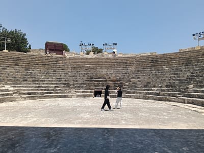 The image shows an outdoor amphitheater with stone seating that curves around a central stage area. The amphitheater appears ancient, constructed from weathered stone blocks. In the foreground, a dark, flat surface, possibly a stage extension or floor, is visible.

On the stage, two figures are standing. One person, dressed in black, is walking to the left, while another, also in black and white attire, is standing and appears to be adjusting something on their head. Further back, near the entrance to a tunnel-like opening in the seating, a third person is visible, seemingly looking out.

Above the amphitheater, the sky is a clear, bright blue, indicating a sunny day. Several large stage lights on tall metal structures are visible at the top of the seating area, suggesting preparations or a recent event. There are also some trees and a small, reddish-brown structure to the left of the amphitheater. The overall impression is of a historical site, possibly being used for performances or events, under a clear, sunny sky. The location is identified as Episkopi, Cyprus.