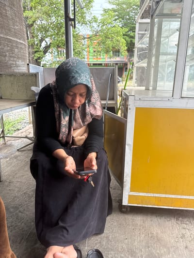 A woman is seated outdoors on a concrete surface, looking down at her mobile phone, which she holds in her hands. She is wearing a black long-sleeved shirt, a dark-colored long skirt, and a patterned headscarf. A tan-colored pouch is visible around her waist. Keys are attached to her phone. To the right of the woman is a yellow and silver food cart. In the background, there are trees and buildings. The scene appears to be a street or outdoor market area in Meunasahmanyet, Indonesia.