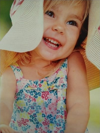 A close-up photograph taken in Sucina, Spain, depicts a young child from the chest up, positioned slightly off-center. The child has fair skin, light blonde hair, and brown eyes, looking upwards and to the right of the frame. The child is smiling widely, exposing a full set of primary teeth. A light-colored straw hat, possibly white or cream with a faint red stripe pattern on the brim's underside, partially covers the child's head and face, with the brim obscuring part of the left cheek and the top of the forehead. The child is wearing a sleeveless top or dress featuring a light background with a multi-colored floral pattern in shades of pink, red, blue, green, and yellow. A horizontal ruffle or bow detail is visible across the upper chest area of the garment. The background is uniformly out-of-focus, presenting a soft green color, suggesting an outdoor setting. No other individuals or specific objects are identifiable in the blurred background.