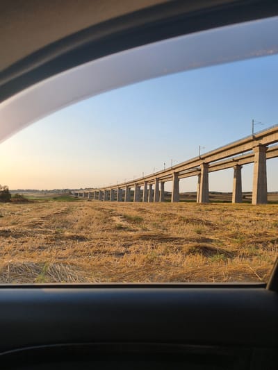 The image presents an exterior view captured from within a vehicle, partially framed by the dark gray upper and lower window sills. The scene depicts a rural landscape during late afternoon or early evening, characterized by warm, golden light. A clear blue sky occupies the upper portion, gradually lightening to a pale orange-yellow near the horizon. The foreground outside the vehicle features an extensive field of harvested crops, predominantly golden-brown stubble and scattered straw, with isolated patches of green vegetation. Stretching across the midground is a long, multi-span concrete railway viaduct, supported by numerous tall, uniformly spaced concrete piers. The viaduct carries railway tracks equipped with an overhead catenary system for electric trains. In the distant background, to the left of the viaduct, a cluster of trees and some green foliage are visible, along with a small, indistinct settlement or village. The horizon line suggests gently rolling terrain. No individuals or specific actions are discernible. The location is the Mate Yehuda Regional Council, Israel.