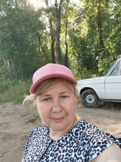A woman wearing a pink baseball cap and a leopard-print top is standing in front of a white car. The car is an older model, possibly from the Soviet era, with a spare tire mounted on the side. The background consists of trees and foliage, suggesting an outdoor setting. The lighting indicates it is daytime, possibly late afternoon, with the sun filtering through the trees. The ground appears to be dirt or sand. The location is identified as Chkalov, Russia, though no specific city landmarks are visible. The woman is looking towards the camera, and her expression is neutral.