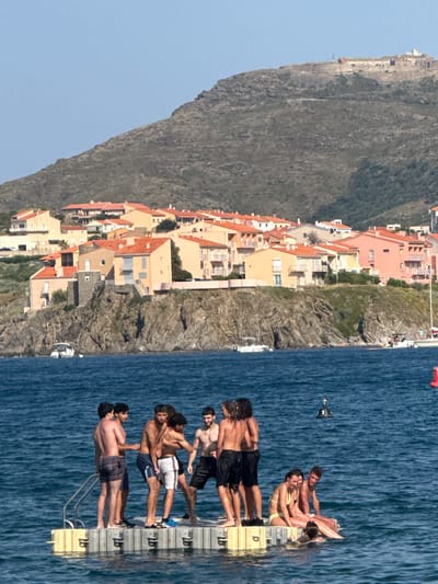 A group of young people are on a floating platform in the blue water near the coast of Collioure, France. The platform has a yellow edge and metal railings. On the platform, several young men are standing and interacting, with some appearing to be pushing or wrestling each other. Other people, including young women, are sitting on the platform. Some are wearing swimwear. In the background, there is a rocky coastline with a town of light-colored buildings with orange tile roofs nestled on the slopes. Above the town, a large, barren hill rises, topped with a historic structure that appears to be a fort or castle. The sky is clear and blue. The water is calm, with a few small buoys visible.