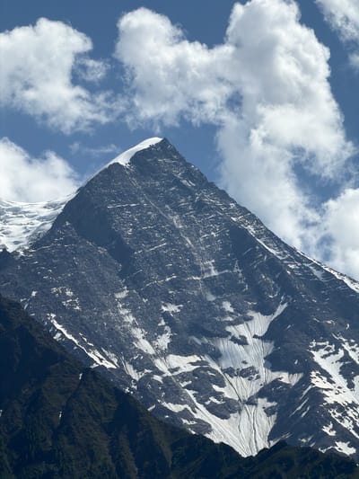A close-up, low-angle shot captures the imposing peak of a snow-capped mountain under a bright blue sky dotted with fluffy white clouds. The mountain's face is predominantly dark rock, sharply contrasted by patches of white snow that cling to its slopes in jagged patterns. Glacial ice is visible on the upper left portion of the mountain. The foreground shows a steep, dark green mountainside, possibly covered in dense vegetation. The scene conveys a sense of ruggedness and natural grandeur. The location is identified as Chamonix-Mont-Blanc, France.