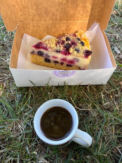 The image displays an outdoor scene in Tomé, Chile, featuring a single slice of berry crumble cheesecake and a cup of black coffee resting directly on green and dry grass. The cheesecake slice is contained within an open, white cardboard box with a brown interior lid. The dessert exhibits a base layer of yellow cake, a creamy white filling, and a topping composed of mixed red and blue berries, covered with a yellow streusel crumble. It is placed on white parchment paper inside the box. A round pink sticker, printed with "Gracias GRACIAS OBRIBA" in white text, is visible on the exterior front of the box. A white ceramic mug with faint red and green patterns, containing a dark brown liquid, is positioned on the grass in front of the cheesecake box. The scene is illuminated by natural daylight.