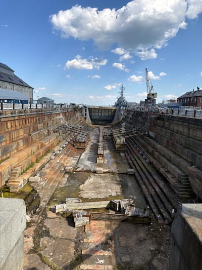The image depicts a dry dock in Boston, United States of America. The dry dock is a large, rectangular structure with stepped sides. The walls of the dock are made of concrete and stone. The bottom of the dock is concrete. There are several large ships in the dock, and a crane. The sky is blue with white clouds. Some buildings are visible on the upper edges of the dry dock.