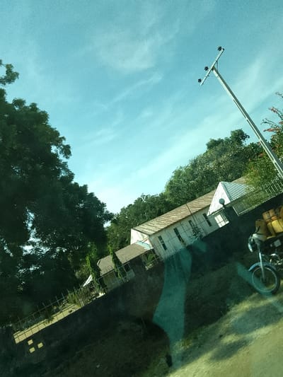 The image depicts an outdoor scene, reportedly in Jibam, Nigeria, under a bright, light blue sky with scattered faint cirrus clouds. The photograph is captured at an angle, exhibiting prominent green and dark reflections across the lower and central areas, consistent with an image taken through a tinted window.
In the midground, a long, single-story building with a light-colored, possibly white, facade and a light-brown corrugated metal roof is visible. The building features multiple rectangular windows. To its left, a smaller, darker-roofed building is partially discernible amidst dense green trees. A portion of a white, pointed roof structure is present to the right of the primary building. A dark, low retaining wall extends along the base of these structures.
On the right side of the frame, a tall, light-colored utility pole with a crossarm supporting four visible insulators stands against the sky. In the lower right foreground, the front section of a light-colored motorcycle is present, displaying a front wheel, fender, and handlebars. Two yellow cylindrical containers, consistent with jerrycans, are affixed to the rear section of the motorcycle, which is mostly out of frame.
Dense green foliage and trees occupy the left side and background of the scene. The immediate foreground consists of an unpaved or dirt surface, partially obscured by the internal reflections. No human subjects are visible in the frame.