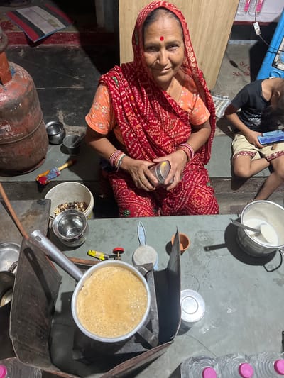 A woman in Pushkar, India, is shown preparing tea in a domestic setting. She is wearing a red and orange patterned sari and a red headscarf. A young boy is seated next to her, looking at a mobile phone. In the foreground, a pot of tea is brewing on a stovetop, surrounded by various utensils and containers, including a gas cylinder, metal bowls, and a bucket with nuts. The scene appears to be an outdoor kitchen area or courtyard.