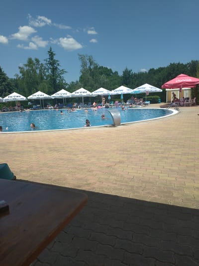 The media file depicts a sunny day at an outdoor swimming pool, likely located in Yagoda, Bulgaria. The scene is vibrant with a clear blue sky dotted with fluffy white clouds. The pool itself is filled with people of various ages enjoying the water. Some are swimming, while others are relaxing or playing. Around the pool, there are lounge chairs and umbrellas, some of which are branded with "DEVIN" and "Coca-Cola," suggesting a commercial establishment like a water park or resort. The poolside is paved with interlocking bricks in shades of yellow and brown. To the right of the pool, a dark shadow suggests the presence of an overhang or structure, and a wooden table occupies the foreground, implying an area for dining or relaxation. The overall atmosphere appears cheerful and relaxed, indicative of a leisure activity on a warm day.