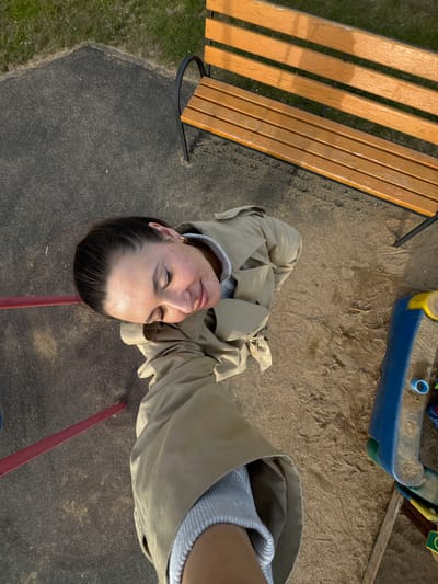 The image, taken from an overhead perspective, shows a woman in a tan trench coat standing in what appears to be a children's playground. A wooden bench is visible on the left, and part of a playground structure, possibly a sandbox, is in the upper right corner. The ground is a mix of pavement and sand. The woman's face is centered in the frame; she has her eyes closed. The location may be Akulovo (Gp Kubinka), Russia, based on provided metadata.