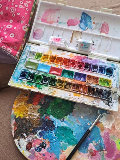 The image shows an overhead view of art supplies, likely for watercolor painting. The primary subjects are a white plastic watercolor paint set and a round artist's palette covered in a variety of mixed paints. The paint set is open, revealing two compartments. The left compartment contains several wells with dried or wet paint residue in shades of pink and blue. Two small tubes of paint are also present in this compartment; one is labeled with text that appears to be in English, and the other has a white label with some Cyrillic lettering and a barcode, which is likely the origin of the location context. The right compartment of the paint set holds rows of individual pans of watercolor paint in various colors, including blues, purples, reds, oranges, yellows, greens, and black. Many of these pans appear to be wet or have been recently used.

The artist's palette, to the right of the paint set, is circular and made of plastic. It is heavily laden with thick, textured blobs of paint in a wide spectrum of colors, including vibrant blues, greens, reds, yellows, pinks, purples, and browns. The paint is applied thickly, suggesting active use. A paintbrush with a black handle and silver ferrule rests diagonally across the palette, its tip touching the mixed paints.

In the lower left corner of the image, a portion of a pink fabric is visible. This fabric has a pattern of white circles with concentric rings and swirling decorative elements. A hint of a purple zippered edge is also visible along the bottom.

The scene is likely set indoors, given the lighting and the presence of art supplies. There are no people visible. The activity taking place is implied to be painting or preparing to paint. The overall impression is one of artistic creation and a used, active workspace. The visible text on the paint tube includes "WATERCOLOR GOUACHE" and "CELESTIAL BRUSH" which can be inferred to be part of the paint's description. The Cyrillic lettering on the other tube, combined with the provided location context of Popovo, Bulgaria, suggests the items may have been purchased or used in Bulgaria.