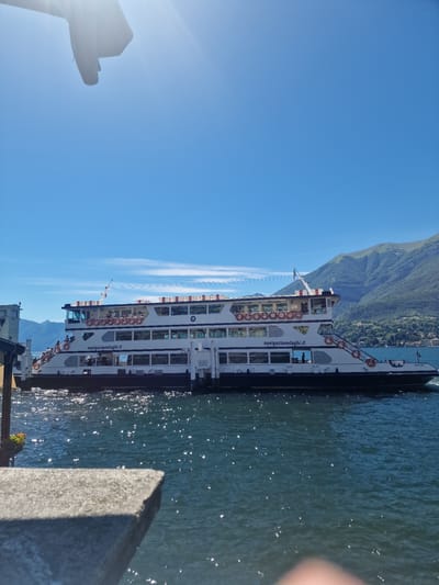 A large, white passenger ferry with multiple decks is docked on a body of water under a bright blue sky. The ferry has "navigazionelaghi.it" written on its side and is adorned with orange life preservers. There are people visible on the decks of the ferry, and some are standing on a ramp leading up to it. In the background, lush green mountains rise, with buildings nestled on their slopes. The water is choppy and reflects the sunlight, creating a shimmering effect. In the foreground, a concrete structure, possibly a pier or wall, is partially visible, with a blurred object in the bottom right corner that may be a person's hand. The scene appears to be in a scenic location, likely a lake or coastal area in Italy, possibly near San Giovanni.