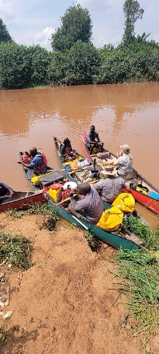 The scene depicts a group of approximately eight individuals in at least five canoes on a reddish-brown, muddy river at Nyabisindu, Tanzania. The canoes, colored red, green, and dark, are positioned along a steep, reddish-brown earthen bank with sparse green vegetation.
In the foreground, a dark green canoe rests partially on the bank, containing two individuals. One, wearing a dark shirt, sits facing forward with a paddle, surrounded by yellow dry bags and other gear, including red fuel canisters. The other is partially obscured. Adjacent to this, a red canoe holds one person wearing a blue life vest. Further into the river, a dark canoe contains one person in a dark shirt and another individual obscured by gear. Another red canoe holds two individuals, one with a dark complexion in a dark shirt, and another light-skinned individual wearing a light-colored shirt and a wide-brimmed hat, looking towards a neighboring canoe. A fifth canoe, partially visible on the far left, contains at least one person.
The canoes are heavily laden with various items including yellow dry bags, red plastic fuel containers, white buckets, paddles, and other packed equipment, indicating a journey. The river's surface reflects the overcast sky, and dense green foliage, comprising trees and bushes, lines the opposite bank under a partly cloudy sky.