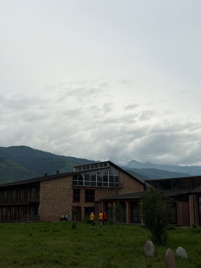 The image depicts a large, multi-story building constructed primarily of brick. The building features numerous windows, including a large, multi-pane section on the upper level, and balconies on the lower levels. A group of people, some wearing yellow shirts, are visible near the entrance. The building is situated in a grassy, outdoor environment with rolling green hills and mountains in the background, under a cloudy sky. Several large stones are placed in the foreground. The location is specified as Shabdan, Kyrgyzstan.