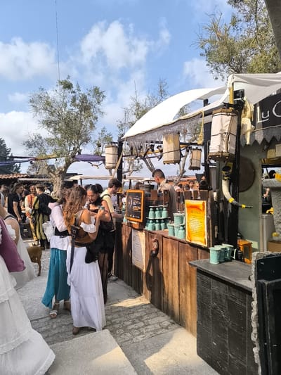The image captures a scene at a medieval fair in Óbidos, Portugal, under a clear blue sky with scattered clouds. A wooden stall, decorated with medieval-style lanterns and chains, is the central focus. Several people are gathered around the stall, some dressed in historical costumes, suggesting a reenactment or festival.

In the foreground, two women in long, flowing dresses are interacting with each other and the stall. One woman on the left wears a white dress with a blue skirt and white sandals, while the woman on the right is in a white tiered dress with black accents and a black shoulder bag. A small dog is visible near their feet.

Behind them, other fairgoers mill about, some in period attire, including a man in a yellow and green tunic. The stall itself is adorned with various items, including mugs, a horn, and a sign with text. The wooden counter displays rows of teal-colored mugs. The background shows more tents and trees, indicative of an outdoor event. The overall atmosphere appears festive and lively.