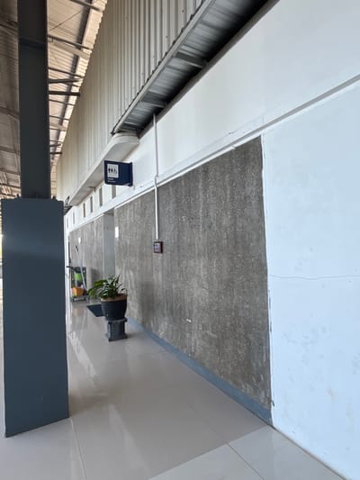 An outdoor hallway with a corrugated metal roof extends to the right, supported by a dark grey pillar on the left. The hallway floor is tiled in a light beige color. The walls on the right are a rough, grey texture with some white paint. A dark blue sign indicating "Toilet" and featuring figures representing men, women, and a person in a wheelchair is mounted on the white wall. Further down the hallway, a cleaning cart with a yellow bucket and mop is visible. A potted plant sits on a pedestal near the cleaning cart. A red fire alarm box is mounted on the grey wall. The scene appears to be in a public area, possibly a transportation hub or a commercial building in Desa Tambakreja, Indonesia, given the architectural style and the tropical foliage.