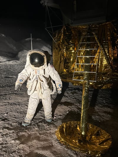 The image depicts a detailed diorama of an astronaut on the moon, set against a backdrop of a mountainous lunar landscape under a dark, star-speckled sky. The astronaut, clad in a white spacesuit with a reflective visor, stands in the foreground. To the right of the astronaut is a golden, foil-covered structure resembling a lunar module, with a ladder attached. The ground is covered in dark, granular material that mimics lunar regolith, showing tire tracks and footprints. The scene is illuminated by artificial lighting, casting shadows on the ground. The environment appears to be a museum exhibit, likely within the city of Salzburg, Austria, given the context provided.