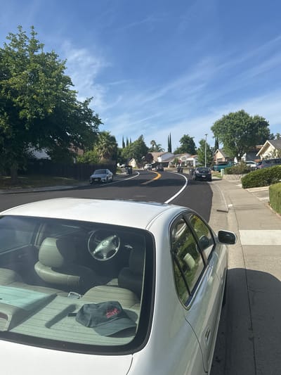 The image shows a residential street in Citrus Heights, USA, on a bright, sunny day. A white car, from which the photo appears to be taken, is prominently featured on the right side of the frame. Its interior is visible through the windshield, revealing cream-colored seats, a steering wheel, and a black hat with red text on the front passenger seat.

The street ahead is newly paved with dark asphalt, clearly marked with a continuous white line and a yellow line on the left side of the lane. A concrete sidewalk runs along the right side of the road. On both sides of the street, houses are nestled among lush green trees and hedges, and several other vehicles are visible further in the distance. The sky is a vibrant blue with sparse, wispy white clouds, indicating clear weather. The strong sunlight casting distinct shadows suggests it is late morning or early afternoon, depicting a typical suburban environment. No people are visible.