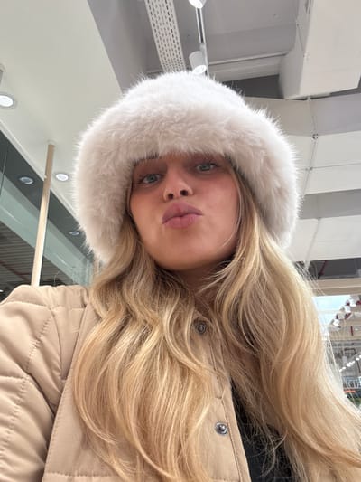 A blonde woman with fair skin is positioned in the center, wearing a white fur-trimmed hood and a tan quilted jacket. She is looking directly at the camera with blue eyes and slightly pursed lips. Her long blonde hair cascades around her face and over her shoulder. The background includes parts of a building with visible lights and structures. The image is rotated slightly.