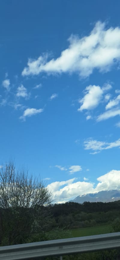 The image depicts a bright blue sky with scattered white clouds. In the periphery, there is a glimpse of mountains with snow. In the lower right of the image, one can see tree branches, green vegetation, and part of what seems to be a metal guardrail. The image provides no information suggesting it was taken in the city of Strumyani, Bulgaria. The picture appears to be taken from a moving vehicle.