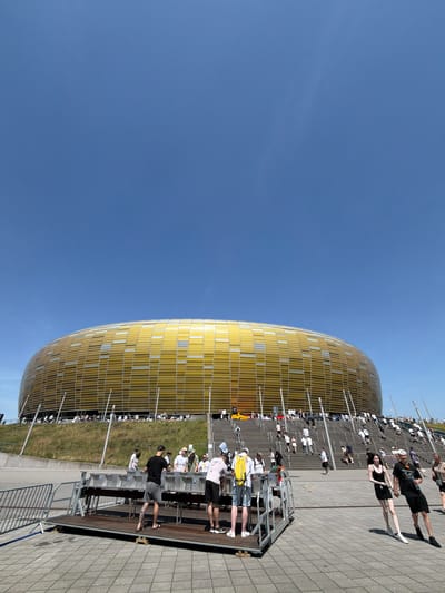 The image depicts the PGE Arena in Gdansk, Poland, a modern stadium with a distinctive golden facade. The sky is a clear, bright blue, indicating a sunny day. In the foreground, a group of people are gathered around a raised platform with several tables, possibly setting up for an event or a market. Some individuals are actively engaged in tasks, while others appear to be observing or socializing.

Behind the platform, a wide set of stairs leads up to the stadium entrance. Numerous people are ascending these stairs, dressed in casual summer attire, suggesting a large gathering or event is taking place. The stadium itself, with its curved, metallic golden exterior, dominates the background, its modern architecture contrasting with the natural green slope to its left. The overall scene suggests a lively outdoor event at a prominent sporting venue.