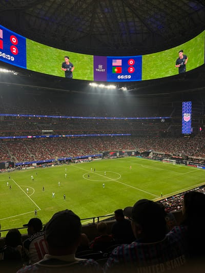 The image captures a live soccer game at Mercedes-Benz Stadium in Atlanta, United States. The stadium appears to be packed with spectators. On a large curved screen overhead, the score is displayed as USA 0 - Portugal 2. The time remaining in the game is 66:59. An image of a soccer player wearing number 18 is also shown on the screen. A young fan in the foreground is wearing a Donovan jersey with the number 10. An individual behind the fan is wearing clothing with a United States design. The soccer field is in view with the game in progress.