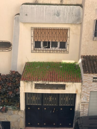 The image displays the exterior facade of a building situated in M'diq, Morocco. The primary structure features a cream-colored stucco finish. At the top, a concrete ledge or parapet is visible, topped with sparse, dry vegetation. Below this, a prominent rectangular window is secured by a black wrought iron grille with an intricate geometric pattern. Behind the grille, a light brown woven blind partially covers the window, beneath which a three-pane window is present; the central pane is noticeably ajar, angled inwards from the bottom.

Beneath the window, a small, sloped canopy or roof is covered with reddish-brown clay tiles, interspersed with neatly arranged parallel lines of vibrant green moss or artificial turf. A narrow, rectangular, screened ventilation opening is located directly beneath this canopy. The ground level features a large, black metal double garage door or gate. This gate is embellished with central gold-colored ornate motifs on each door panel and smaller gold star-shaped or floral accents near the bottom edges; a gold-colored handle is present on the right door. To the left of the garage door, a section of wall with a stone base is partially obscured by a dense bush with dark, reddish-tinged foliage, behind which a black metal fence segment is visible. An adjacent building on the right exhibits a similar tiled roof and a section of wall faced with light-colored brick-patterned tiles, with a portion of a white roll-up garage door visible at the bottom right. No individuals are depicted in the scene.