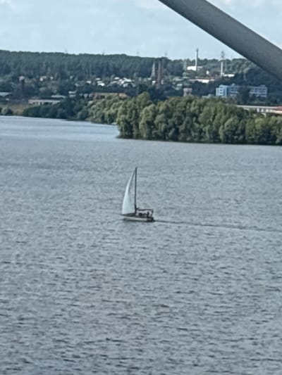 A sailboat with a white sail navigates a wide body of water, possibly a river or lake. The water surface is textured with ripples. In the background, a dense line of green trees borders the far shore. Behind the trees, a city skyline is visible, featuring various buildings, industrial structures, and tall towers. The sky above is overcast and light-colored. A dark, diagonal beam, possibly part of a bridge or overhead structure, cuts across the top of the image. The scene appears to be located in Zelenodol'sk, Russia, given the context of the provided information.
