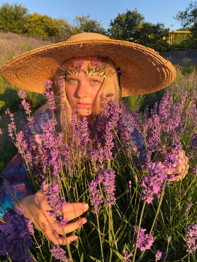A young woman with blonde hair and striking blue eyes lies amongst a field of blooming lavender. She wears a wide-brimmed straw hat adorned with sprigs of lavender, and a glimpse of her face is visible beneath its shadow. Her expression is serene, with her lips slightly parted. The setting is a lavender field, indicated by the abundance of purple flowers and green stalks. The sun is setting, casting a warm, golden light on the scene, suggesting it's late afternoon or early evening. The sky is a clear blue, hinting at fair weather. In the background, trees and a wooden fence can be seen, and beyond that, more rows of lavender stretch into the distance, implying a large cultivation. The woman is partially covered by the lavender, creating a sense of immersion in nature. Her hand rests gently on some of the flower stalks, adding to the peaceful ambiance. The overall mood is tranquil and idyllic. The image was captured in Verkhneye Uchdere, Russia.