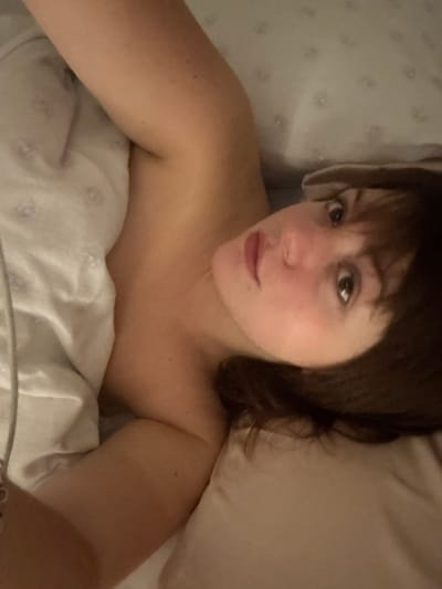 A fair-skinned woman lies in bed, partially covered by white floral-patterned sheets. She faces the camera with a direct gaze, her brown hair framing her face. Her arms are raised above her head. The surrounding bedding appears soft and plush, with a neutral-colored pillow beside her. The scene is casually lit, suggesting a relaxed or intimate moment.