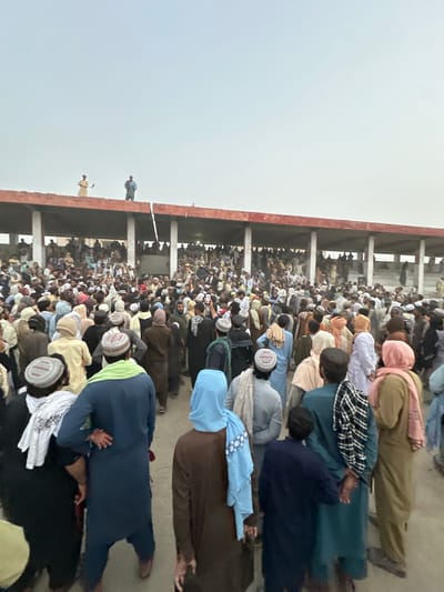The image depicts a large gathering of people, predominantly men, in an open-air area with a covered seating structure in the background. The attire of the individuals suggests a cultural context, with many wearing traditional clothing such as shalwar kameez and turbans or kufi hats. The seating structure appears to be an elevated platform or bleacher-style seating, with columns supporting a roof. Two individuals are visible standing on top of this structure in the distance. The crowd is densely packed, with some individuals appearing to be engaged in conversation or observing something. The environment appears to be a dusty or unpaved outdoor space. The overall scene suggests a public event or gathering, possibly religious or social, in Chaman City Tehsil, Pakistan.