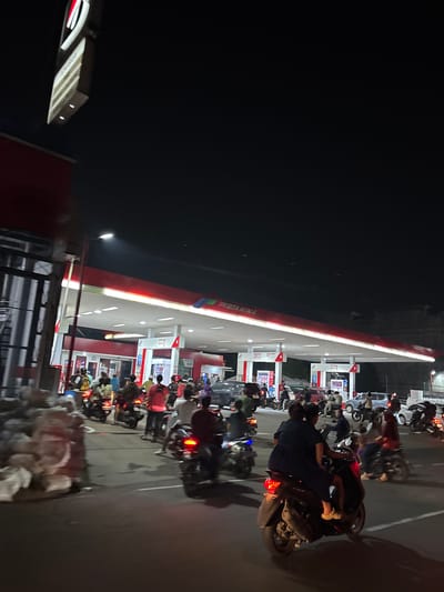 The image depicts a busy night scene at a Pertamina gas station in South Tangerang, Indonesia. A long line of motorcycles and their riders are waiting to refuel. The gas station's canopy is illuminated by fluorescent lights, casting a glow on the red and white structure. The scene is characterized by the presence of numerous motorcycles, with riders dressed in various casual clothing. Some riders are seated on their bikes, while others appear to be dismounted, possibly in conversation or waiting for their turn. The atmosphere suggests a typical evening rush or potentially a shortage of fuel leading to extended queues. The time of day is clearly night, indicated by the darkness of the sky and the artificial lighting. No specific weather conditions are discernible. The word "PERTAMINA" is visible on the gas station canopy, confirming the brand and location. The overall impression is one of a lively, functional hub for motorcycle transportation in an Indonesian urban setting during nighttime hours.