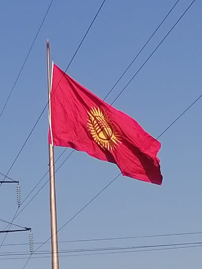 The image shows the flag of Kyrgyzstan flying on a flagpole against a clear blue sky. The flag is red with a yellow emblem in the center. The emblem is a stylized representation of a yurt's crown. The flagpole is metallic and appears to be located outdoors. There are power lines visible in the background, crossing the sky. The setting appears to be a relatively open area, possibly in a town or city. The image is taken from a low angle, looking up at the flag and sky. The lower portion of the image shows parts of power line infrastructure. The location is identified as Medrese, Kyrgyzstan.