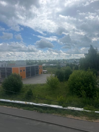 The image depicts an outdoor scene in Kokhma, Russia, under a partially cloudy sky. In the foreground, a paved walkway with a curb runs parallel to a road. Above the walkway, a large, horizontal pipe system, possibly for utilities, runs across the frame. Beyond this, there is a grassy area with various trees and bushes. In the mid-ground, a modern, rectangular building with an orange and dark gray facade stands out. The building features multiple windows. To the left of this building, another longer, light-colored building with numerous windows and the word "ШКОЛА" (School) visible on its facade can be seen. The background is filled with more buildings and trees, suggesting a mixed urban and suburban environment.