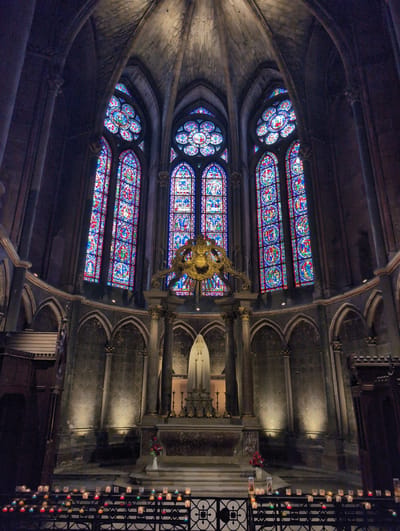 This image depicts the interior of a grand cathedral, likely a place of worship, showcasing its magnificent stained-glass windows and an altar. The cathedral's architecture features high vaulted ceilings and intricate stonework, creating a somber and awe-inspiring atmosphere. In the center, a white statue of a religious figure stands on a raised platform, adorned with flowers and lit by spotlights. In front of the altar, numerous small candles are arranged on a black metal railing, casting a warm, flickering glow. The stained-glass windows, illuminated from behind, display vibrant colors and detailed biblical scenes, adding to the ethereal beauty of the space. There are no people visible in the image, and the scene is static, suggesting a moment of quiet contemplation or prayer. The overall impression is one of reverence and historical grandeur. The provided information states the location is Urvillers, France; however, the architectural style and grandeur of the stained glass windows strongly suggest a much larger and more famous cathedral, such as Notre Dame de Paris, which is a more fitting depiction of such a magnificent structure.