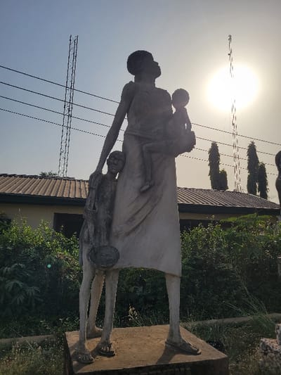 The image presents a weathered concrete statue depicting three figures, identified as the "Monumento a la Familia Negra" or "Monumento a la Esclavitud," located in San Martín, Meta, Colombia. The central figure is a tall, slender adult female standing upright, her head tilted upwards and to the right. She cradles a small child in her right arm, while another slender child figure stands leaning against her left leg. The standing child holds a shallow bowl or plate in their left hand, positioned by their side. All figures exhibit a lean, elongated form.
The statue rests on a rectangular concrete base, surrounded by sparse, dry ground vegetation in the foreground. In the mid-ground, behind the monument, dense green foliage and bushes are visible, partially obscuring a building with a dark, ridged roof and light-colored walls. The background consists of a bright, clear sky, with the sun appearing as an overexposed luminous disc in the upper right quadrant, casting strong backlighting. Two tall, metal lattice communication towers are prominent against the sky, one to the left and another to the right of the sun. Utility lines are visible traversing the upper left portion of the sky. Several slender, tall trees are present on the right side of the background, partially silhouetted.