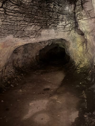 The image depicts the interior of a cave or tunnel, likely man-made, in Lukovit, Bulgaria. The walls and ceiling are rough and textured, composed of rock and possibly some mud or clay. The lighting is dim, with a flashlight beam illuminating parts of the cave, creating shadows and highlighting the uneven surfaces. The floor is uneven, covered in dirt, small rocks, and what appears to be some dampness or shallow puddles. The passage narrows and recedes into darkness in the distance, suggesting a longer, unexplored section. There are no people or discernible objects other than the natural geological formations and the ground cover. The environment is dark, enclosed, and suggestive of an underground natural or historical site.