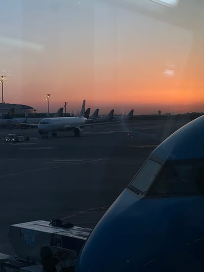 The image depicts an airport tarmac at dusk, viewed through a window. In the foreground, the nose of a blue airplane is visible, with the partial word "XM" on its fuselage. To the left, a cargo container with "KLM" branding is loaded onto a vehicle. In the midground, several airplanes are parked on the tarmac. One prominent white airplane in the center has "AIRFRANCE" written on its side. Other aircraft with visible tail fins are also present. Airport lighting poles with multiple lights are visible in the distance. The background features a building with a curved roof and a city skyline against a sky transitioning from light blue to a vibrant orange sunset. The overall scene suggests operations at an airport, possibly Barcelona-El Prat Airport, located in El Prat de Llobregat, Spain, during the early evening hours.