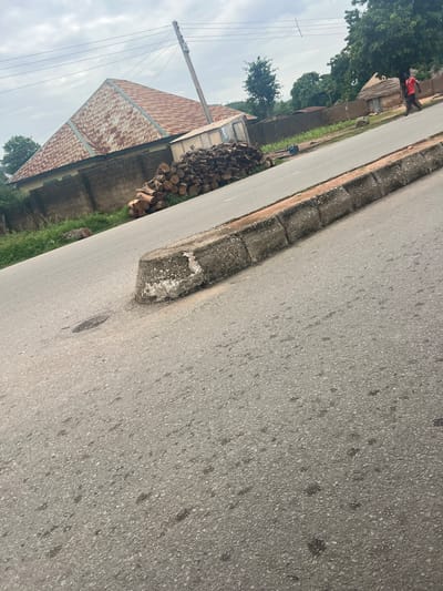 This media file depicts a street scene in Kona Garu, Nigeria. The image is oriented vertically, with the road and its curb taking up a significant portion of the frame. The road surface is asphalt, with visible aggregate and some darker stains. The curb is made of concrete blocks and appears weathered.
To the left of the road, there is a grassy area and a cluster of cut logs, suggesting a recent logging activity or firewood preparation. Further to the left, a building with a corrugated metal roof, showing signs of rust, is visible. Adjacent to this building, there's a smaller structure with a thatched roof. A person wearing a red shirt is standing near the thatched-roof building. Power lines and a wooden utility pole are also present, indicating the presence of electricity. The sky is overcast, suggesting cloudy weather, and the lighting is diffused, implying it might be daytime but not necessarily bright sunlight. The overall scene conveys a rural or semi-rural atmosphere typical of some areas in Nigeria. No visible text is present in the image.